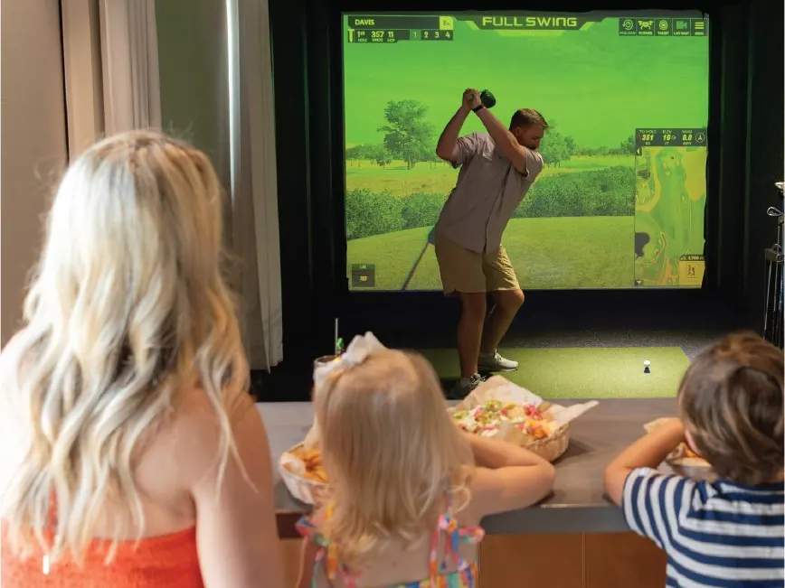 A golfer mid-swing in a Swing Suite bay at Tee Off at The Wharf while a family watches from the counter