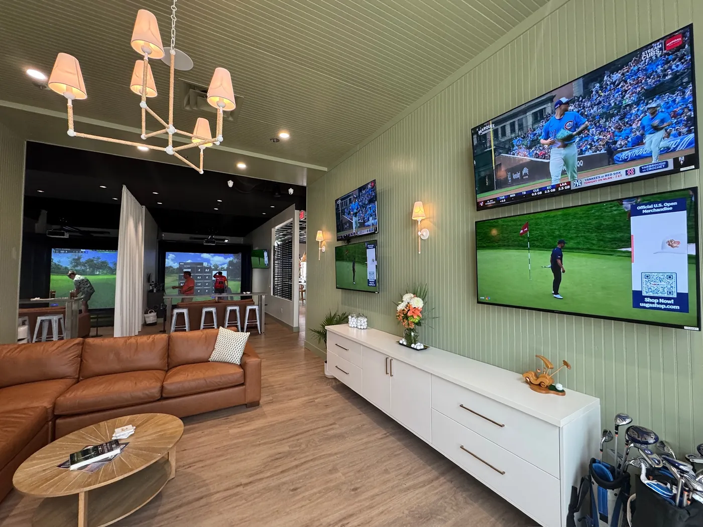 The lounge at Tee Off at The Wharf with baseball and golf on the wall of screens, a leather sectional in front and the simulator bays beyond