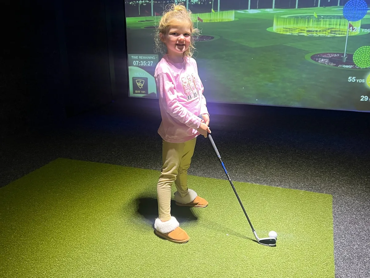 A small child lining up a shot with a club on the turf of a bay at Tee Off at The Wharf