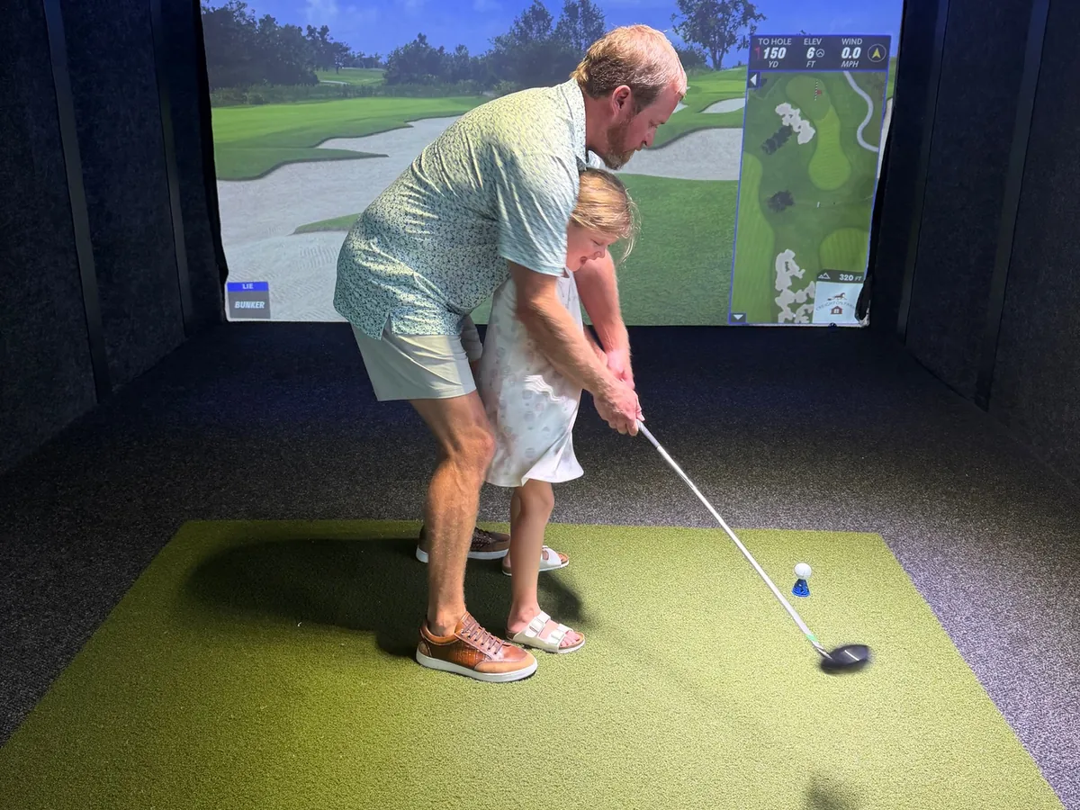 An adult guiding a toddler through a golf swing in a bay at Tee Off at The Wharf