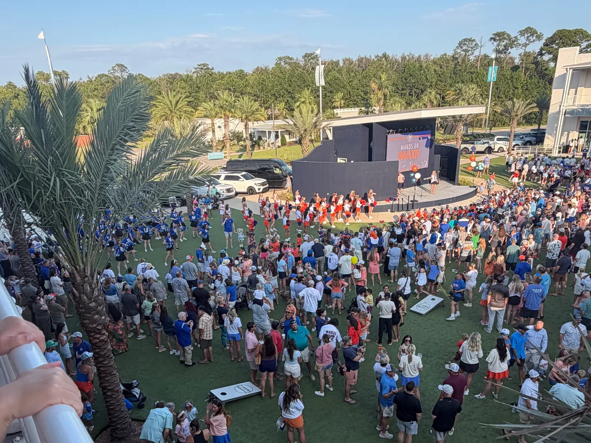 A packed lawn at The Wharf greenspace facing the outdoor stage and its screen, with cornhole boards set up among the crowd