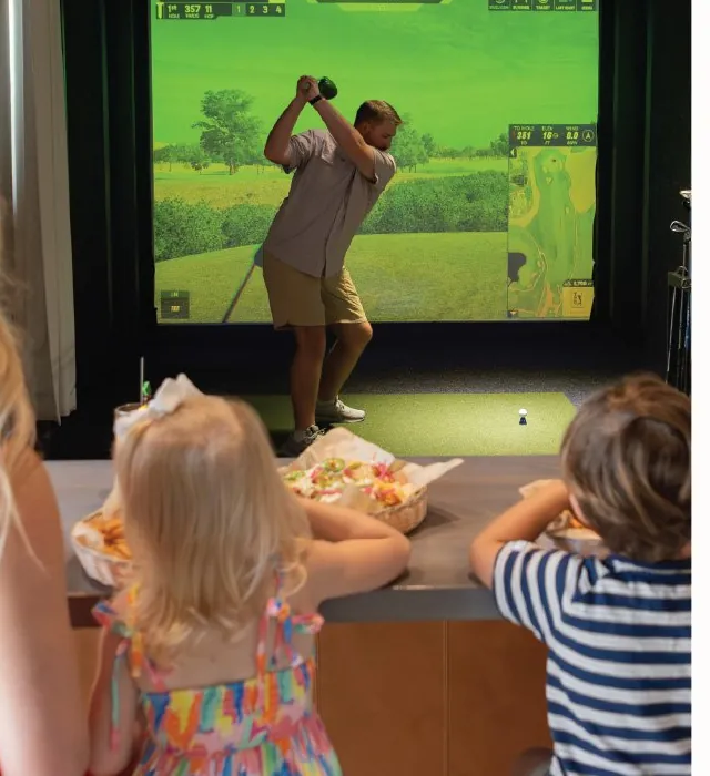 A family watching a golfer swing in a simulator bay at Tee Off at The Wharf