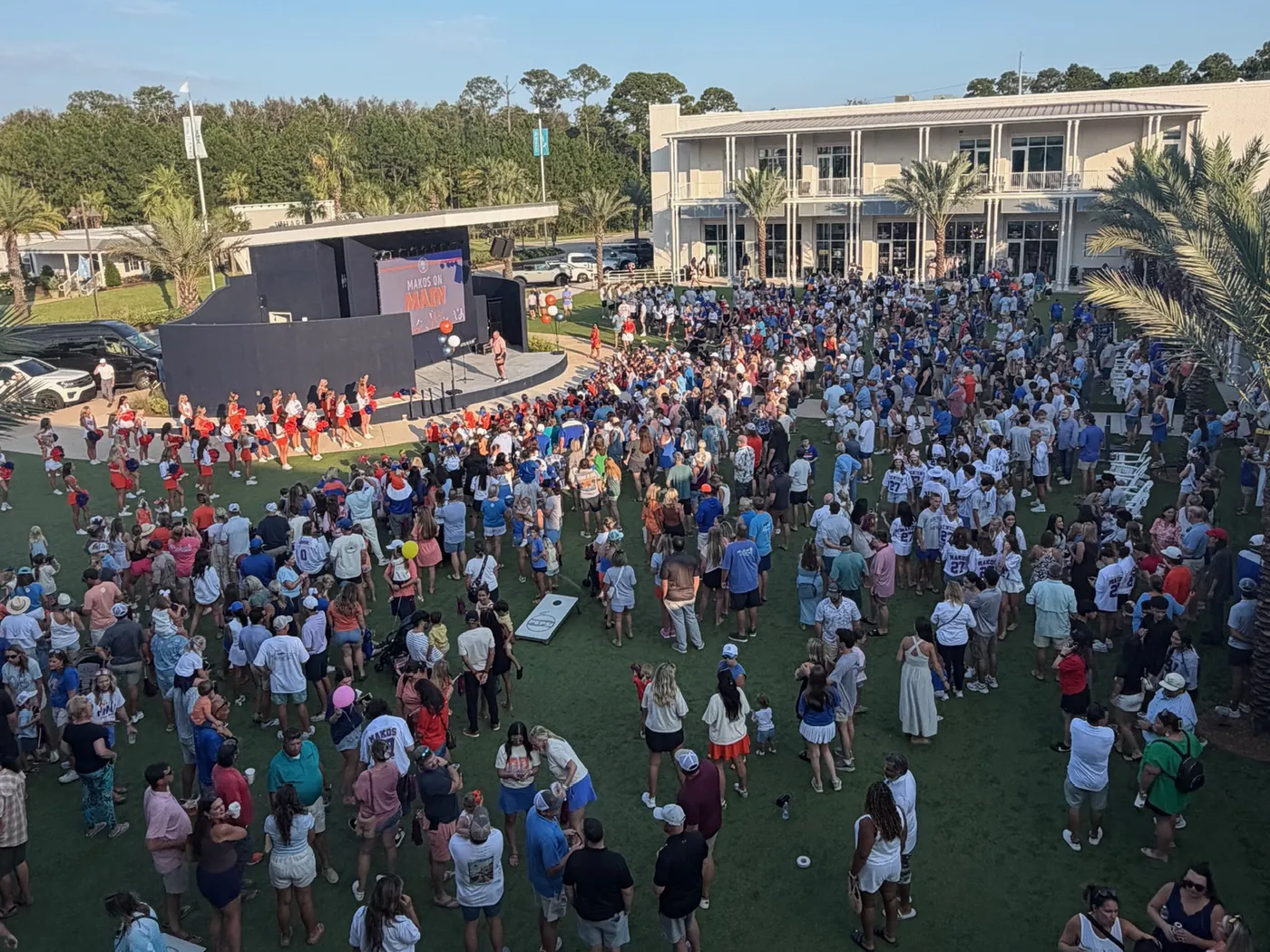 A crowd filling the lawn beside the outdoor stage at Tee Off at The Wharf for a community event, with cheerleaders on the stage and cornhole boards set up on the grass