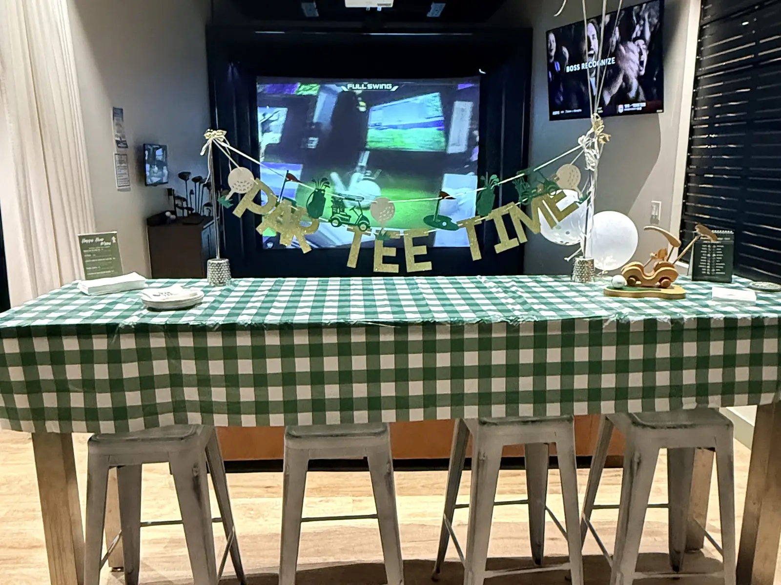 A bay set up for a party at Tee Off at The Wharf — gingham table, balloons and a Tee Time banner under the screens