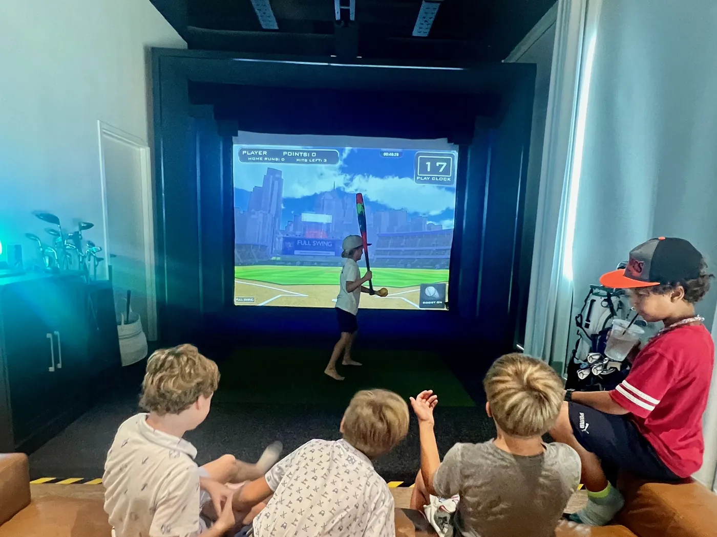A batter at the plate on the screen of a bay at Tee Off at The Wharf, watched from the sofa behind