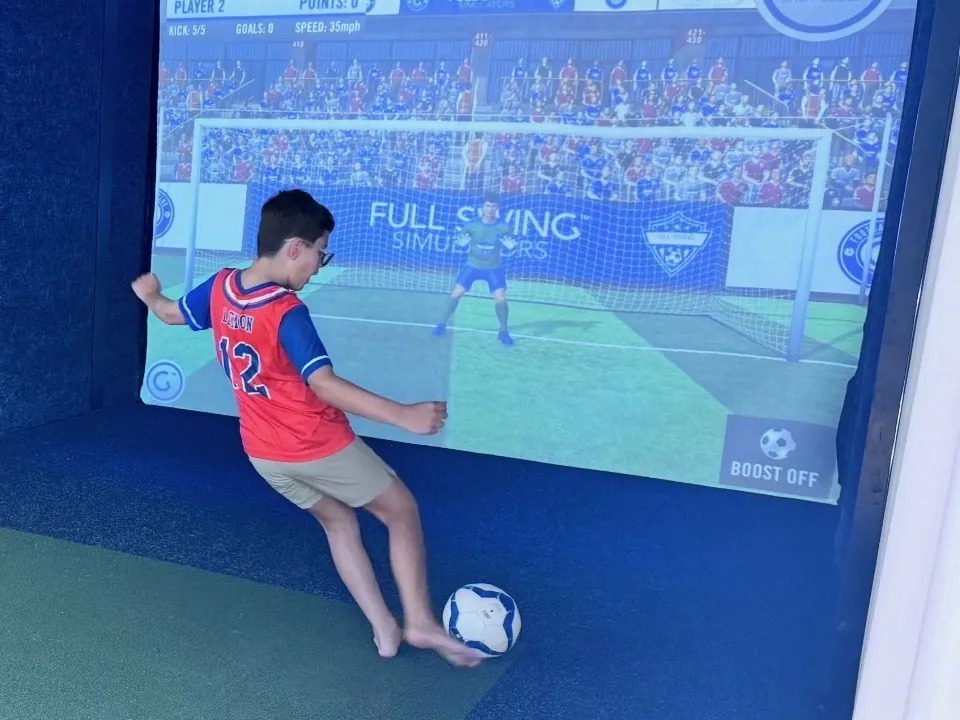 A young player in a soccer shirt striking a ball at the goal on the screen of a bay at Tee Off at The Wharf